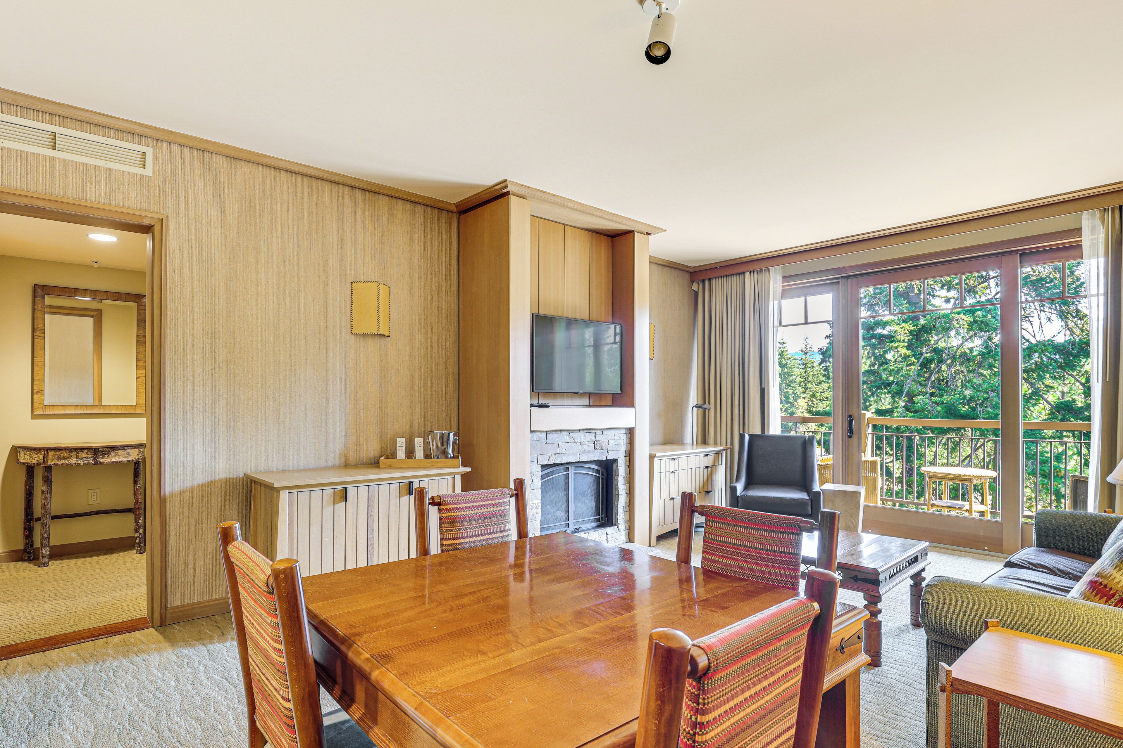 A spacious dining area with a wooden table and chairs, a television mounted above a fireplace, and a balcony with a view of trees.