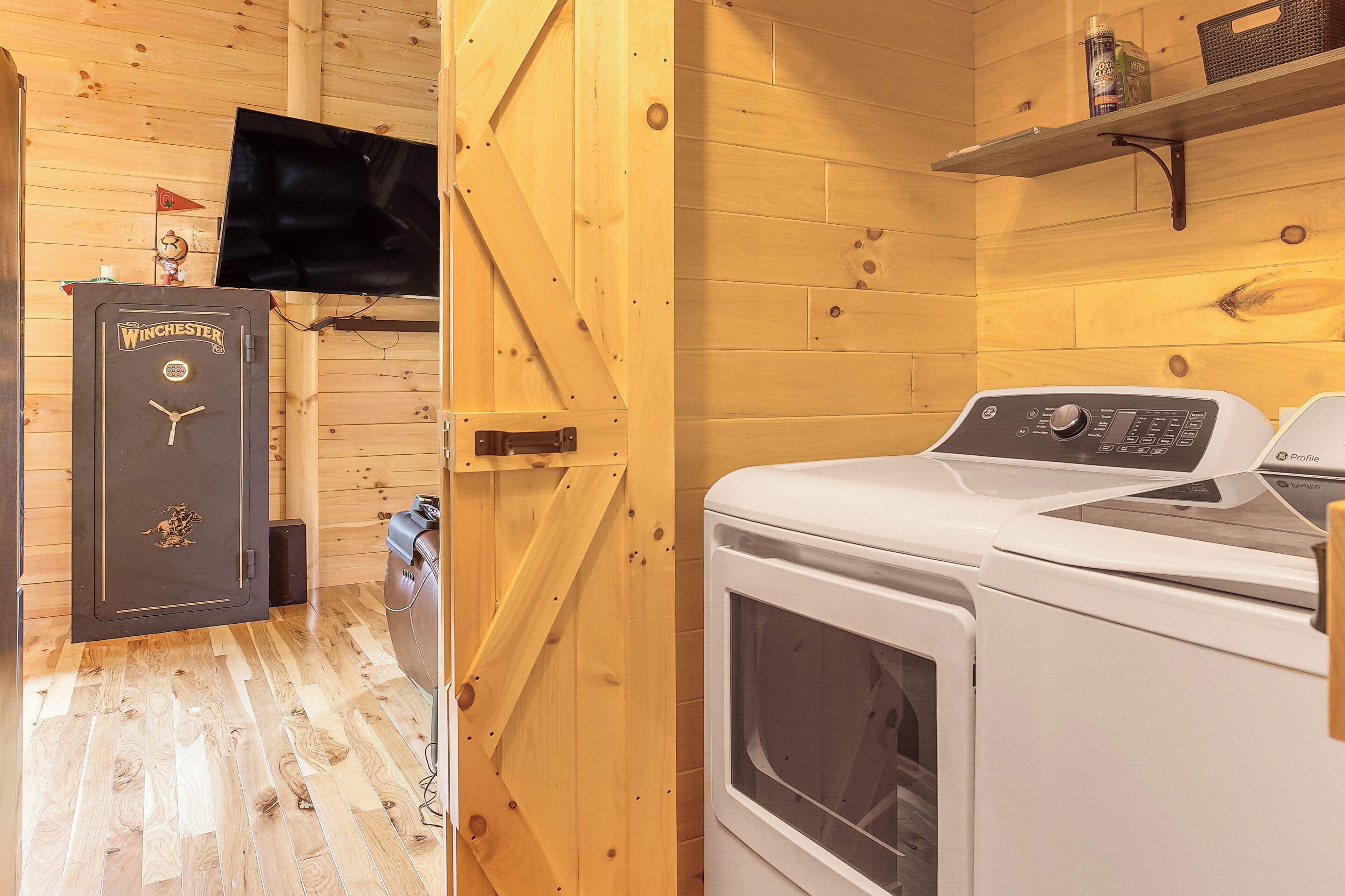 Une buanderie équipée d’un lave-linge et d’un sèche-linge, avec des murs en bois, une étagère sur laquelle se trouve une boîte, et une porte ornée d’un logo.