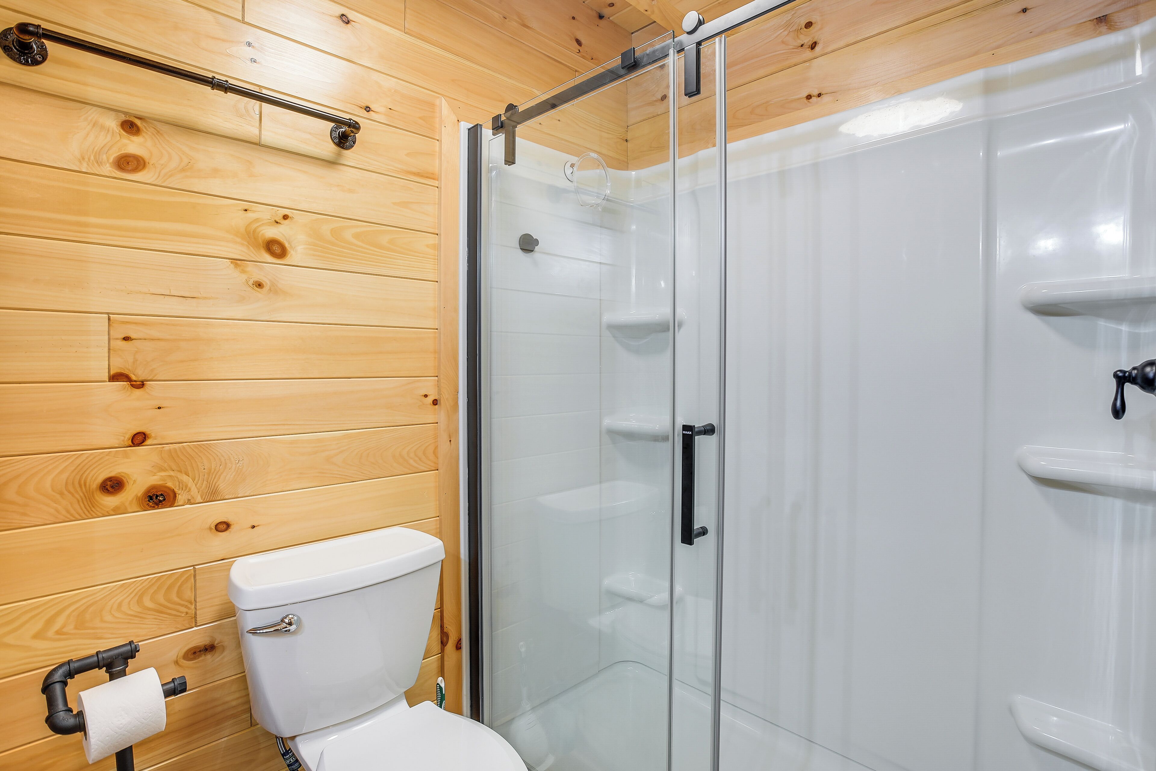 A wooden-paneled bathroom with a glass shower enclosure, a toilet, and a towel dispenser.