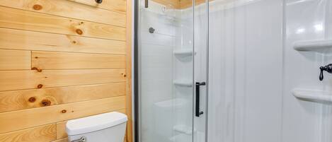 A wooden-paneled bathroom with a glass shower enclosure, a toilet, and a towel dispenser.