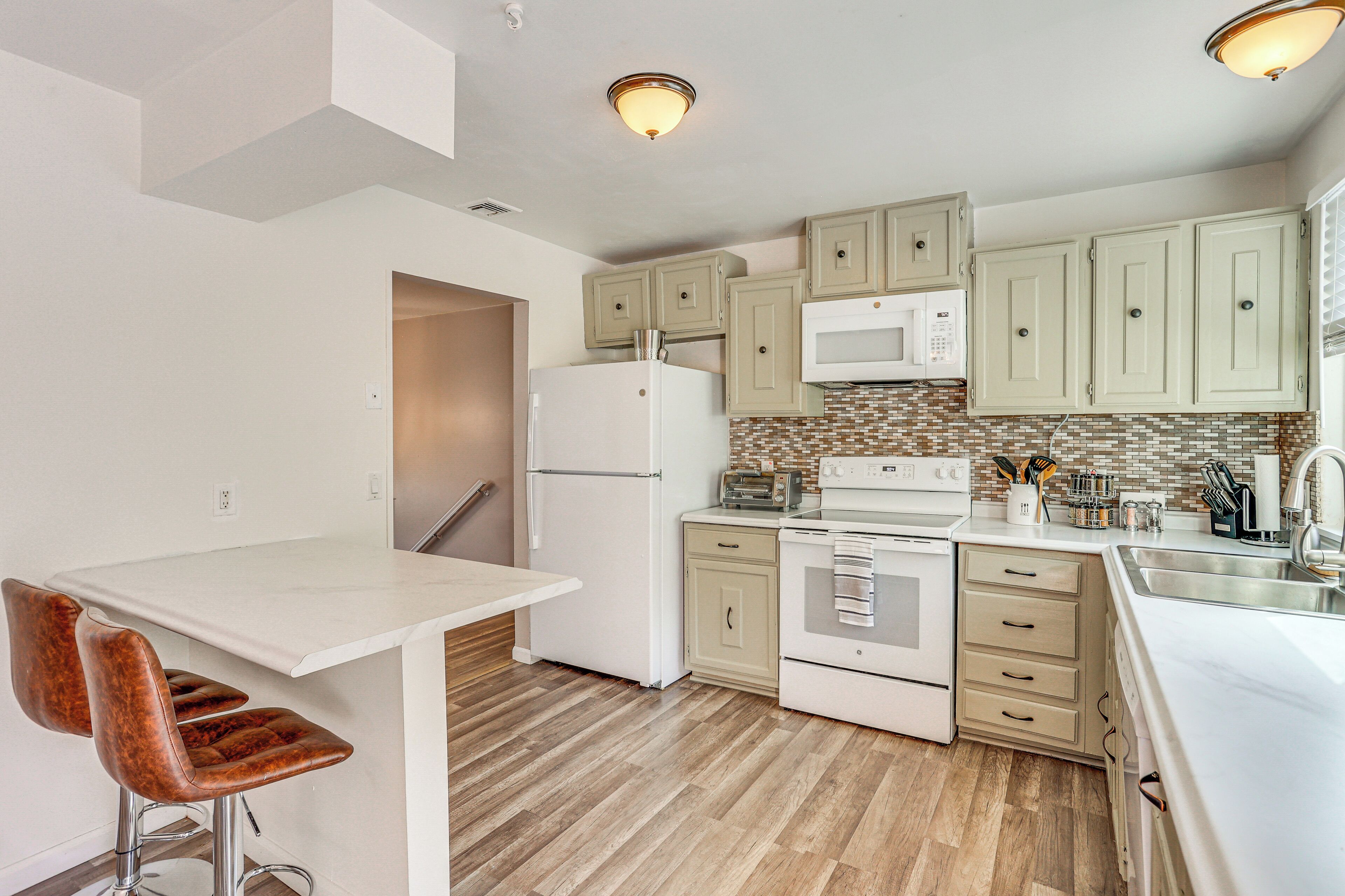 A modern kitchen with white cabinets, a refrigerator, an oven, and a microwave. There is a small island with a sink and a chair.