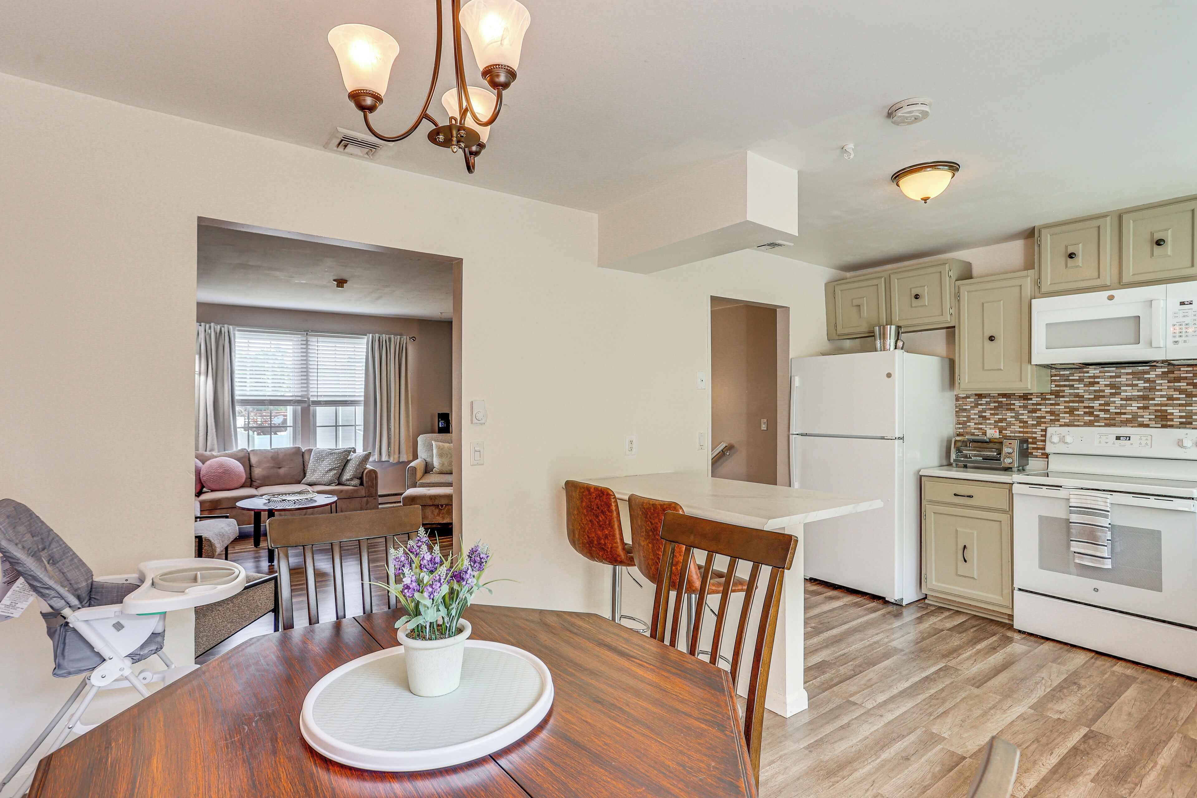 A modern kitchen with wooden cabinets, a white refrigerator, and a microwave. There is a dining table with chairs and a small potted plant.