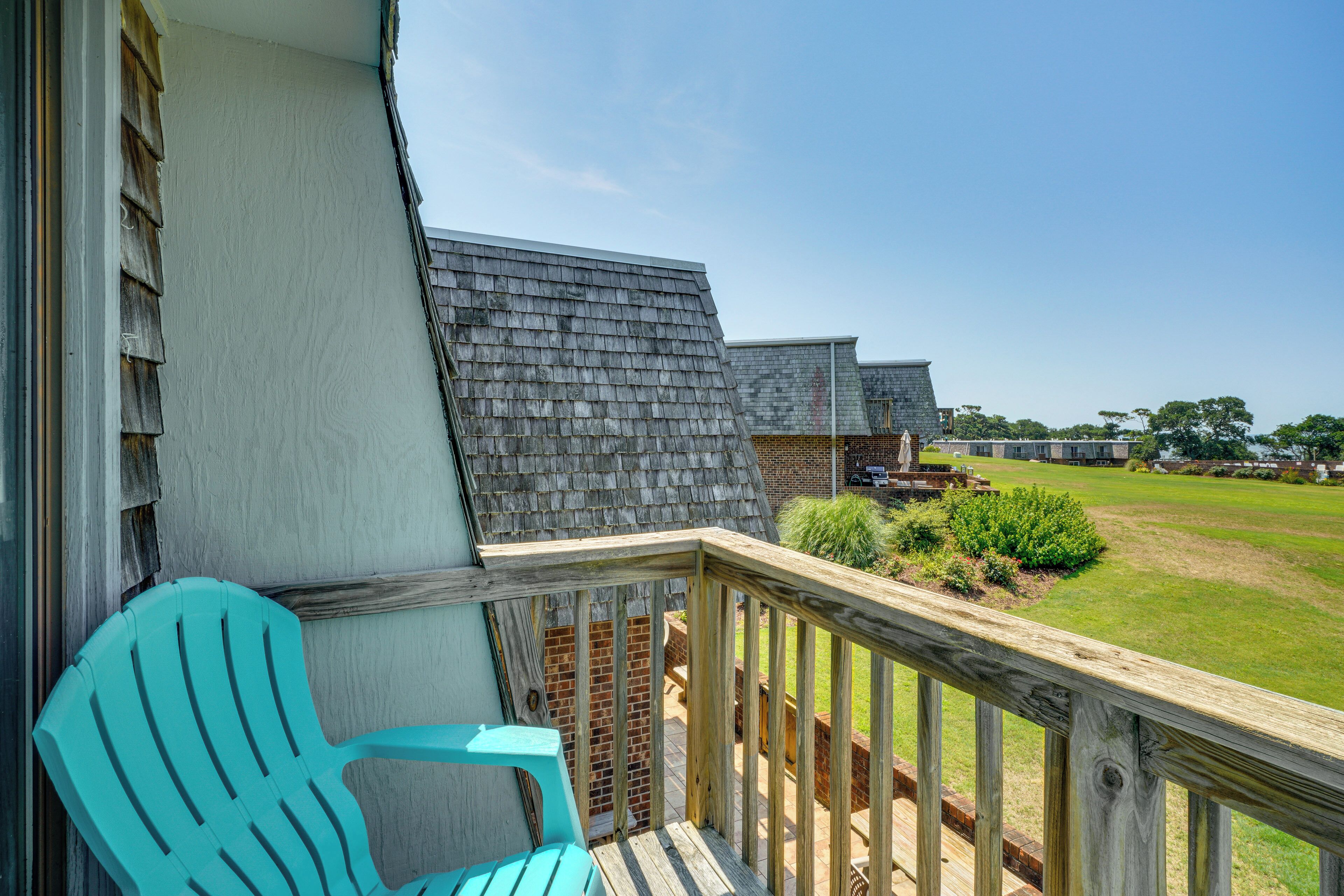 Un balcon doté d’une balustrade en bois, une chaise Adirondack turquoise et une vue sur un espace verdoyant avec des bâtiments en arrière-plan.