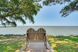 A wooden Adirondack chair on a wooden deck by a lake with a dock in the background. - Waterfront Nags Head Townhome w/ Amenity Access! (Nags Head)