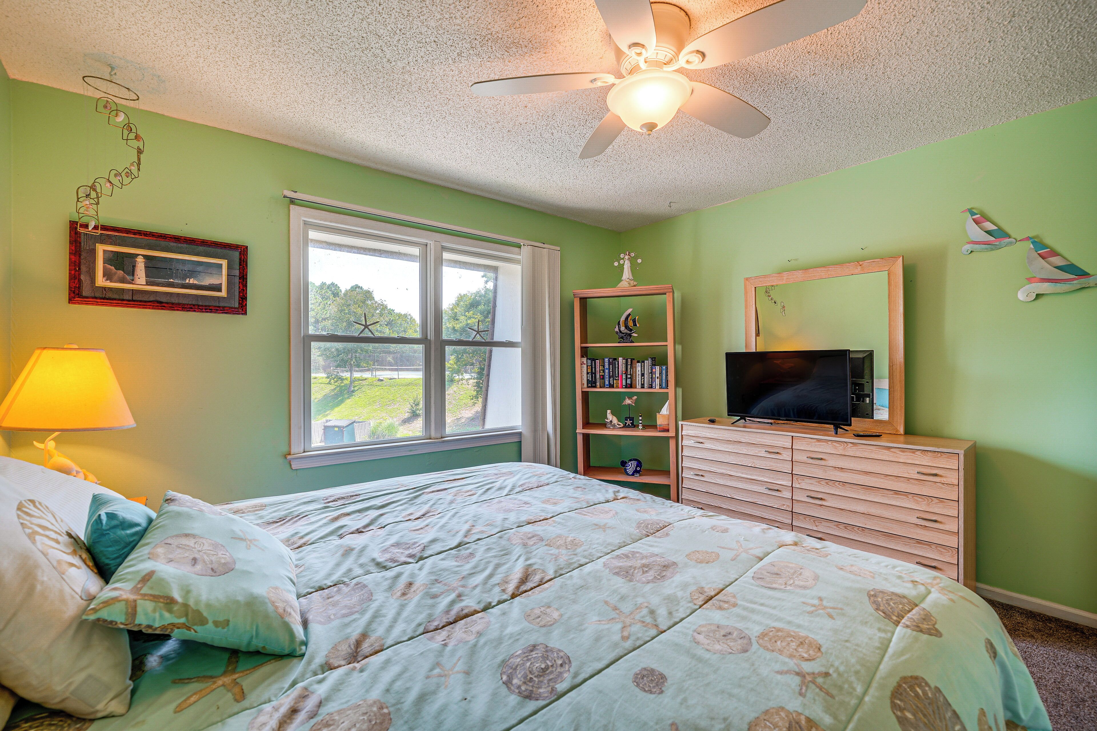 A bedroom with a bed, dresser, television, and a window with a view of greenery.