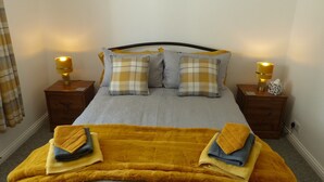 Een net opgemaakt bed met een grijs en geel dekbedovertrek, twee bijpassende kussens en een gele plaid. Er staan twee nachtkastjes met lampen en een zakdoekendoosje op één.