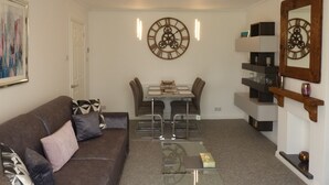 Een woonkamer met een bruine bank, een glazen salontafel, een eettafel met stoelen en een grote wandklok.