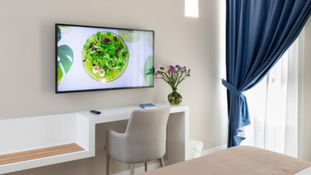 A modern hotel room with a flat-screen TV mounted on the wall, a desk with a chair, a vase with flowers, and a large window with blue curtains.