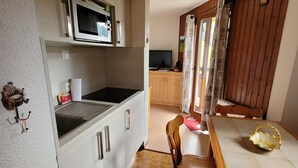 A compact kitchen with a microwave, stove, and wooden cabinets. A dining area with a chair and table is visible. - 2-room apartment for 4 people, pets and parking (Châtel)