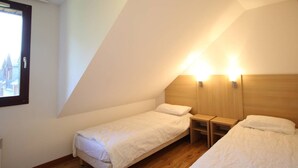 A small, single-room hotel room with two single beds, a wooden headboard, a nightstand, and a window with a view of buildings.