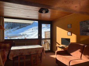 A cozy room with a wooden ceiling, a large window overlooking a snowy mountain, a sofa, a table with a floral tablecloth, and a TV on a wooden stand.