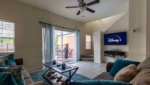 Una sala de estar con televisión, mesa de centro y sofá. Hay una puerta corrediza de vidrio que da a un patio con mobiliario exterior.