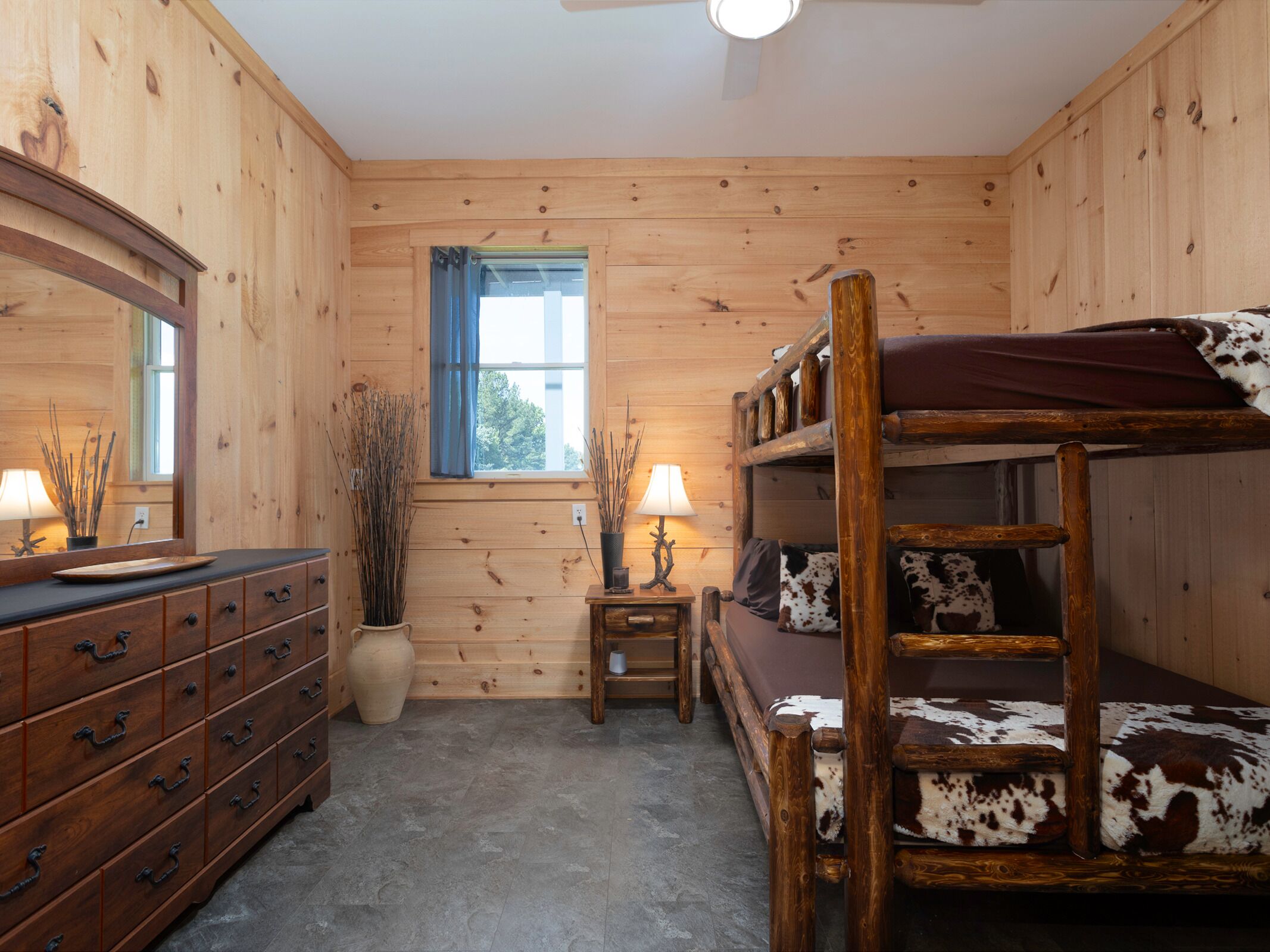 A wooden cabin room with a bunk bed, dresser, mirror, window, lamp, and decorative plants.