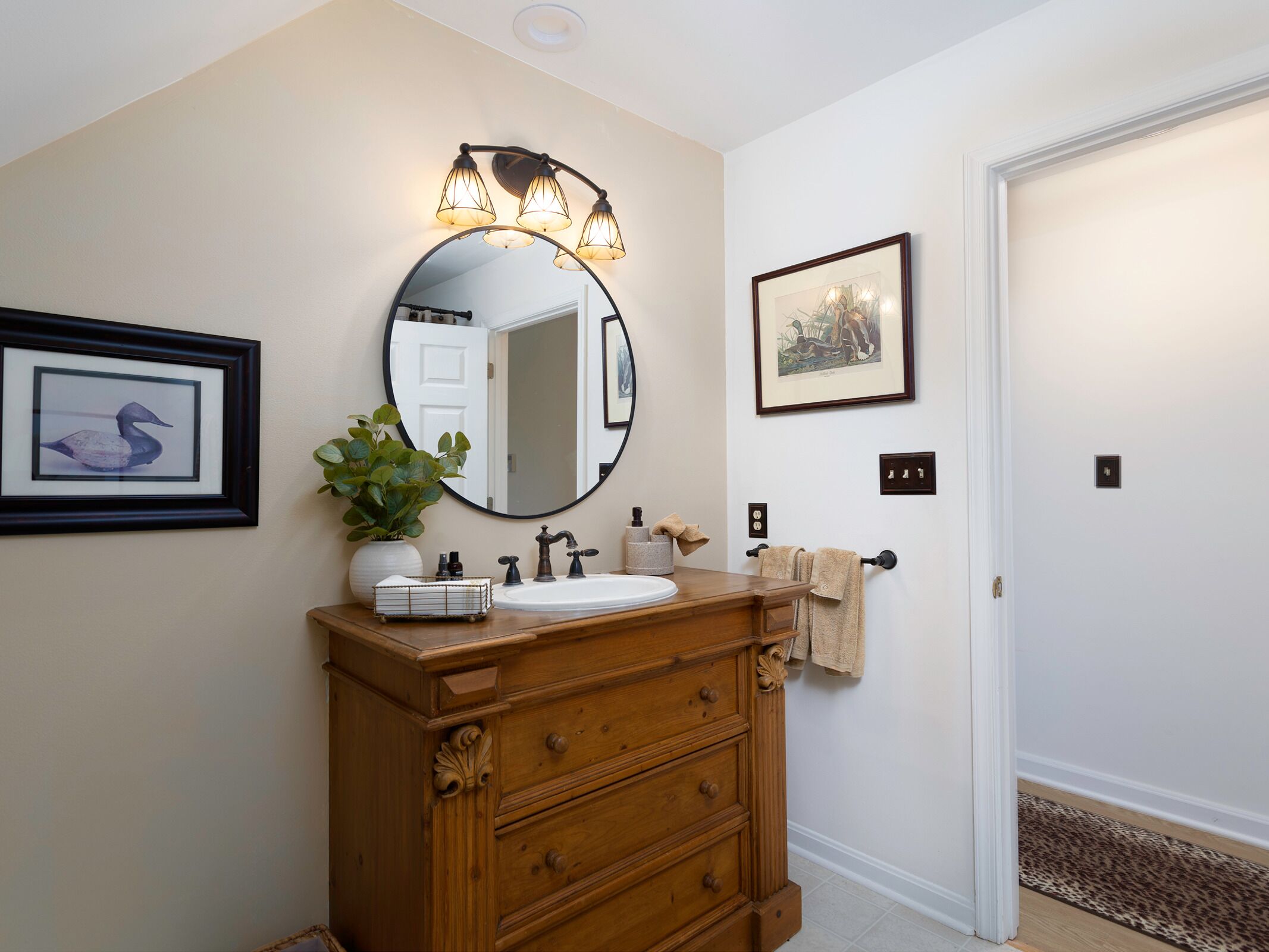 A bathroom with a wooden vanity, a round mirror with lights, a framed picture, and a door with a patterned doormat.