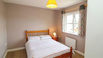 Un dormitorio con cama de madera, mesita de noche con lámpara, ventana con cortinas y radiador.