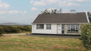 A single-story house with a white exterior and dark roof tiles, surrounded by a grassy yard and a hilly landscape in the background. - THE ISLAND'S VIEW, pet friendly in Gortahork, County Donegal (LETTERKENNY)