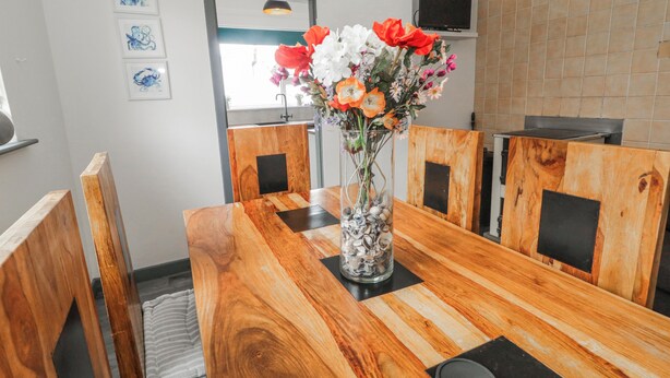 A dining table with a vase of flowers, wooden chairs, a television on the wall, and a tiled backsplash. - THE ISLAND'S VIEW, pet friendly in Gortahork, County Donegal (LETTERKENNY)