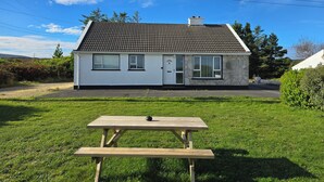 A single-story house with a tiled roof, a front door, and two windows. In front of the house is a wooden picnic table. - THE ISLAND'S VIEW, pet friendly in Gortahork, County Donegal (LETTERKENNY)