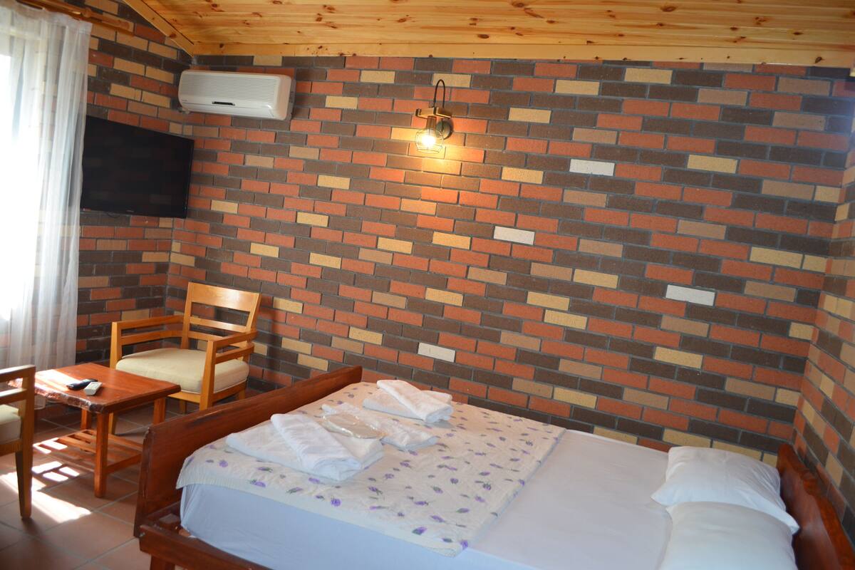 Una habitación con pared de ladrillo, una cama con sábanas blancas, una silla de madera y un televisor de pantalla plana montado en la pared.