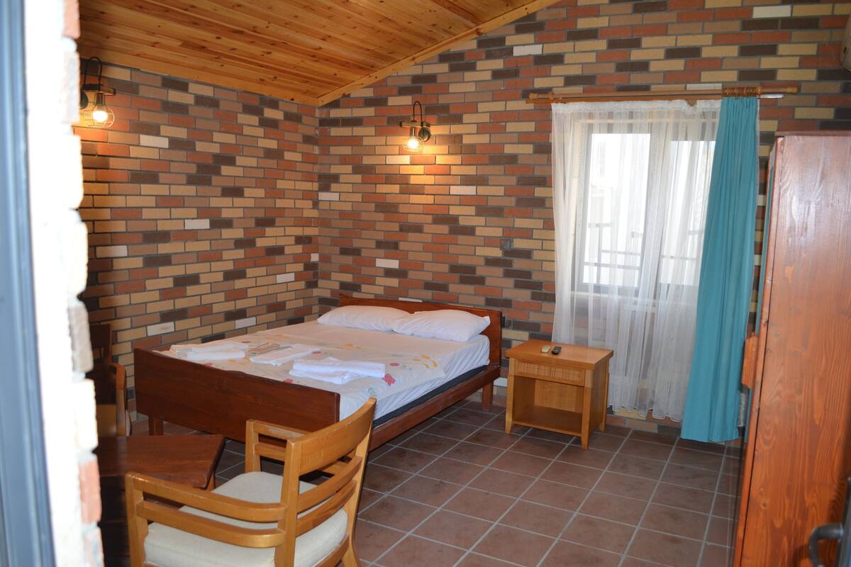 Un dormitorio con pared de ladrillo, cama de madera, mesita de noche, una silla y una ventana con cortinas.