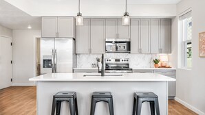 A modern kitchen with grey cabinets, stainless steel appliances, and a white island with three stools. - Paradise Cove 2 Bedroom Condo (St. George)