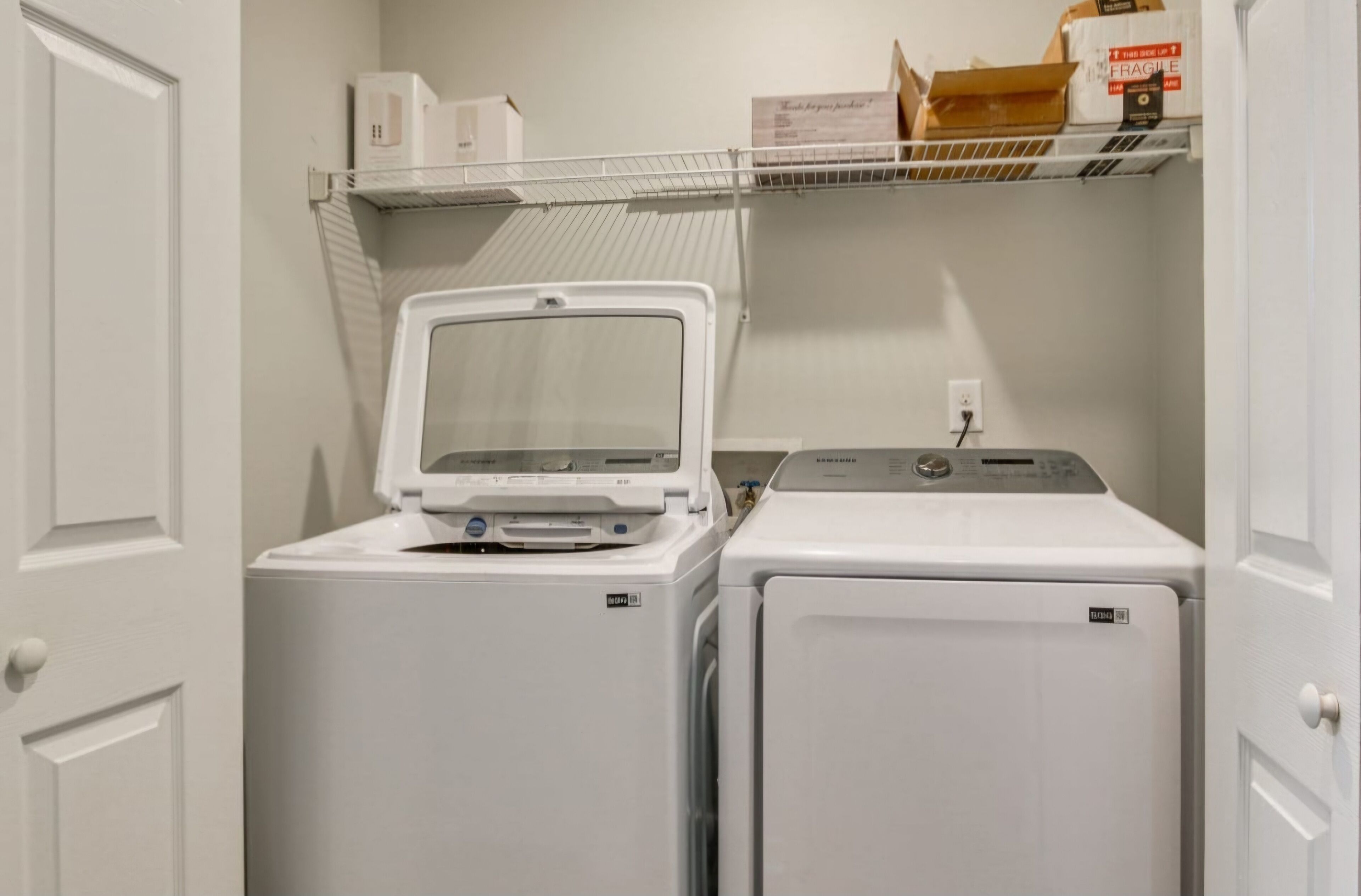 A laundry area with a front-loading washer and dryer, a shelf above with boxes, and a door on the left.