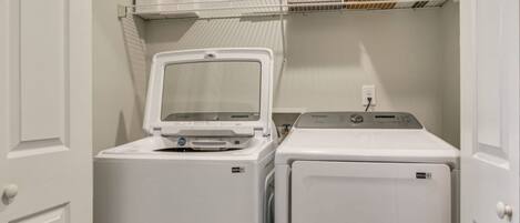 A laundry area with a front-loading washer and dryer, a shelf above with boxes, and a door on the left.