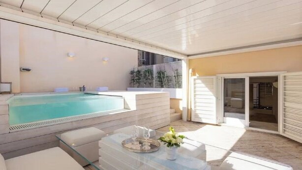 Un área de piscina exterior moderna con una mesa de vidrio, sillas de descanso y una puerta que da a un patio cubierto.