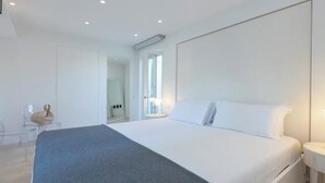 Una habitación de hotel moderna con una cama grande, una mesita de noche con lámpara y vista al pasillo.