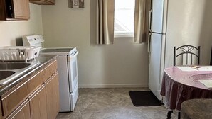 Cocina compacta con pileta, estufa y heladera. Hay una ventana con cortinas, una mesa pequeña con sillas y un reloj en la pared.