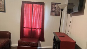 Un salón con sofá, alfombra roja, una pequeña mesa con mantel rojo, televisor en la pared y ventana con cortinas rojas.