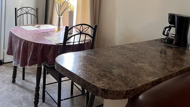 Un comedor con una mesa cubierta con mantel rojo, dos sillas, un jarrón con flores, una cafetera y un refrigerador al fondo.