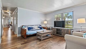 Ein geräumiges Wohnzimmer mit Sofa, Couchtisch und Fernseher, Holzboden und Blick auf Palmen durch das Fenster.