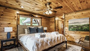 A wooden cabin bedroom with a bed, nightstands, a ceiling fan, and a painting on the wall. - Lookout Lodge- Blue Ridge GA (Blue Ridge)