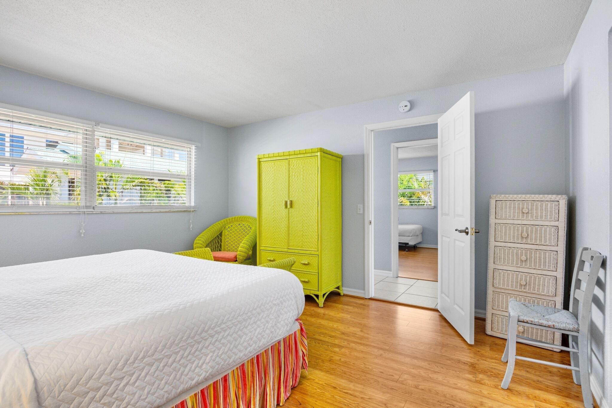 A bedroom with a bed, a green wardrobe, a wicker chair, and a white chest of drawers.