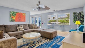 A living room with a sectional sofa, a round coffee table, a ceiling fan, and a view of the outdoors through large windows. - Emerald Shores East! Walk to Beach! (Anna Maria)