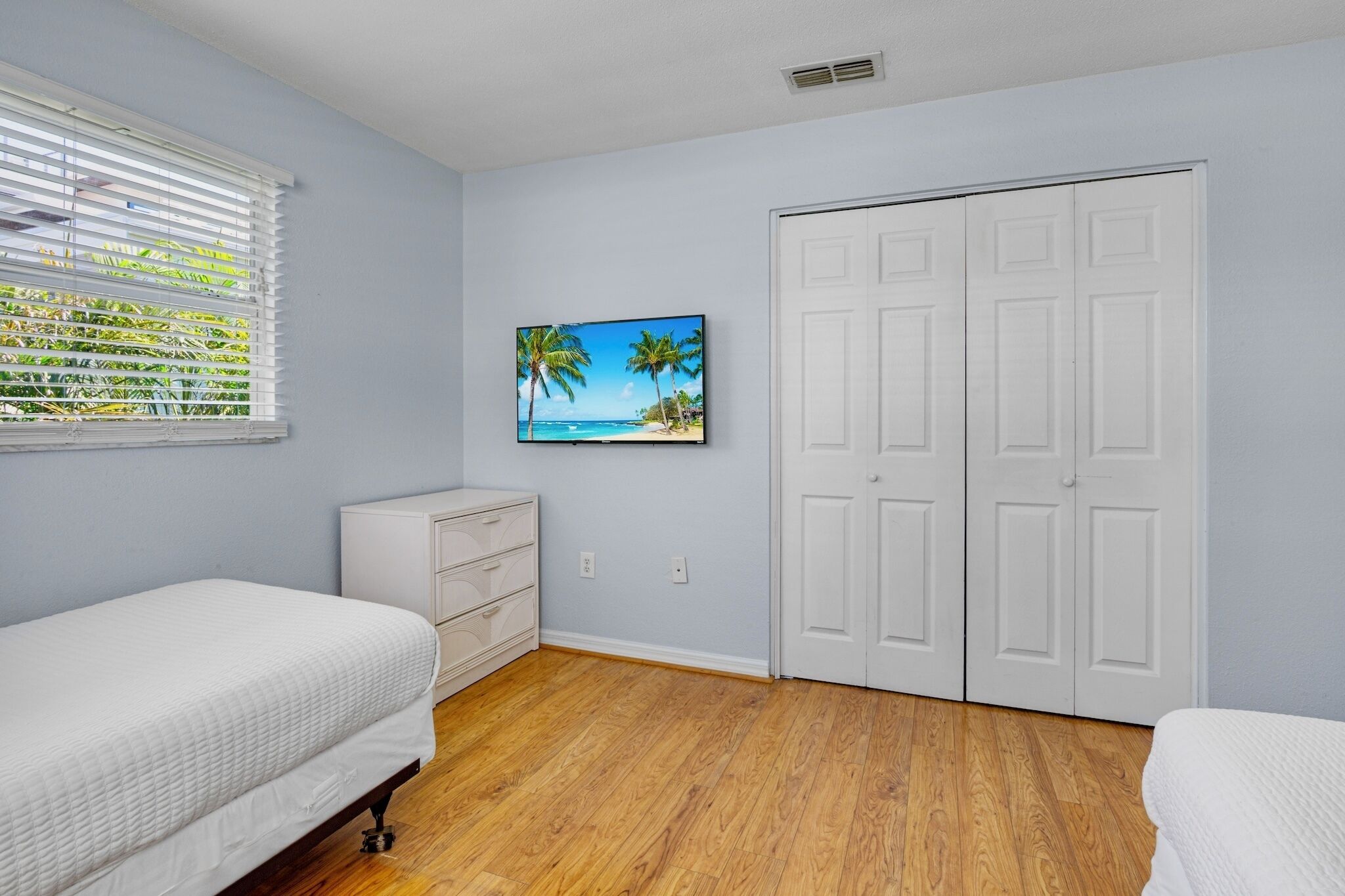 Ein Schlafzimmer mit Bett, Kommode, Schrank und einem an der Wand montierten Fernseher, der eine Strandansicht zeigt.
