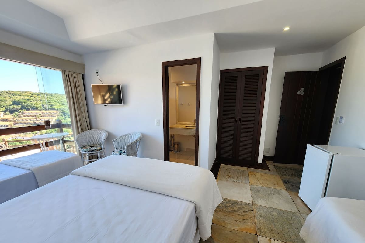 A hotel room with a bed, a TV, a small table, two chairs, a wardrobe, and a view of the landscape.