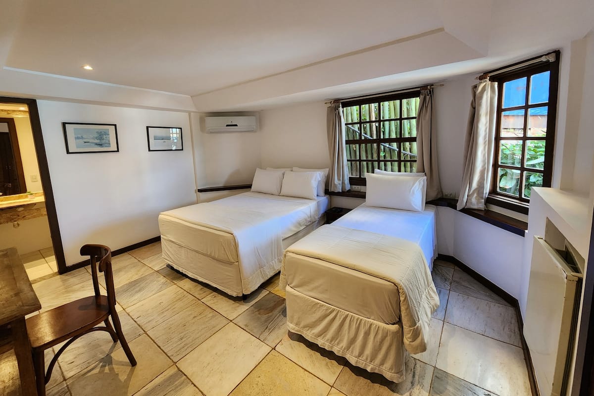 A hotel room with two beds, a desk, and a chair. There are two framed pictures on the wall, an air conditioning unit above the beds, and a window with curtains.