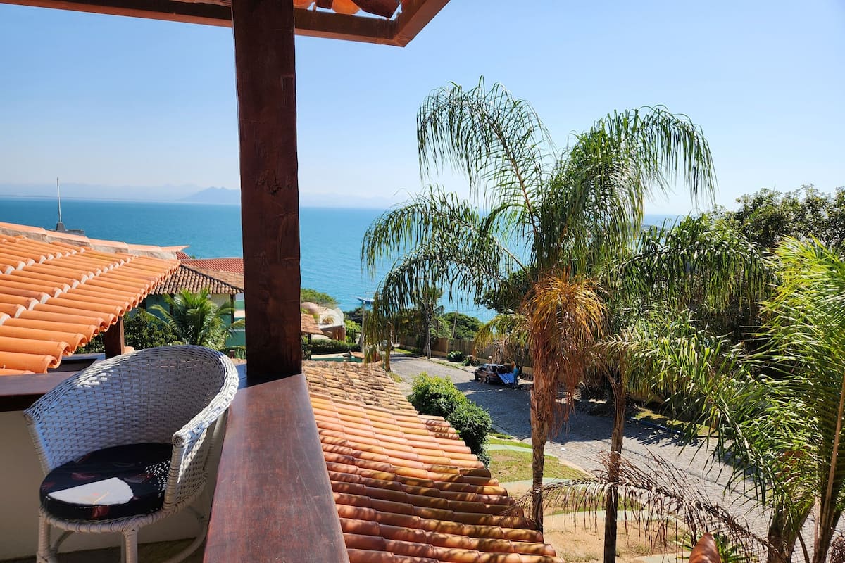 A balcony with a view of the sea, a wicker chair, and a tiled roof.