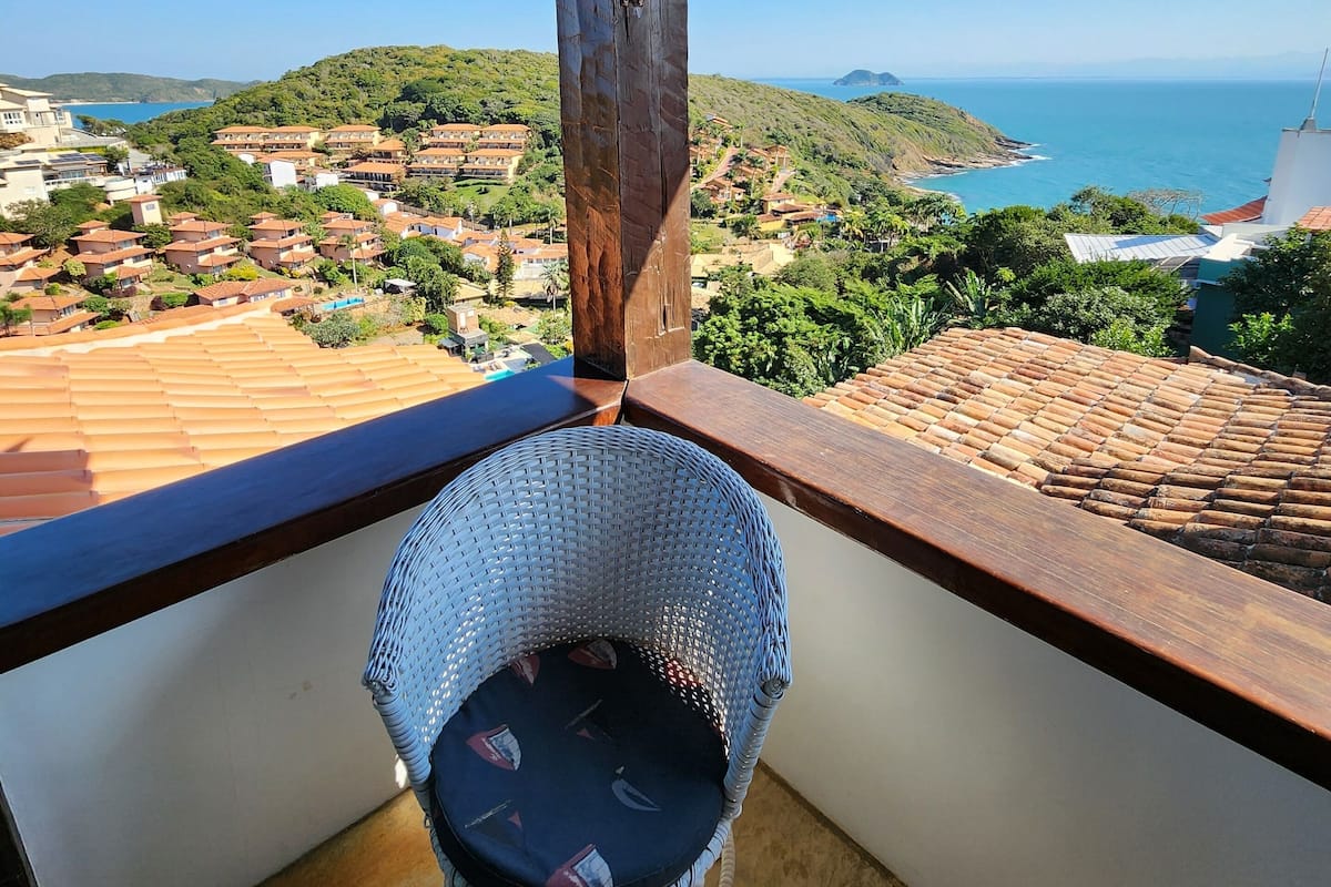 A balcony with a wicker chair, a view of a coastal town, and a wooden ceiling structure.