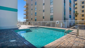 Une piscine avec une rambarde en métal, une terrasse en briques et, au fond, un bâtiment doté de plusieurs balcons.