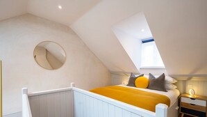 Un dormitorio con techo inclinado, un espejo redondo, una cama con manta amarilla y una mesita de noche con lámpara.