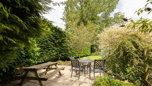 A patio with a wooden table and chairs, surrounded by lush greenery and a well-maintained lawn. - Host Stay Moorland Cottage (York)