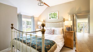 A bedroom with a white metal bed frame, a large window with curtains, a bedside table with a lamp, and a wall-mounted plant. - Host Stay Byre Cottage (York)