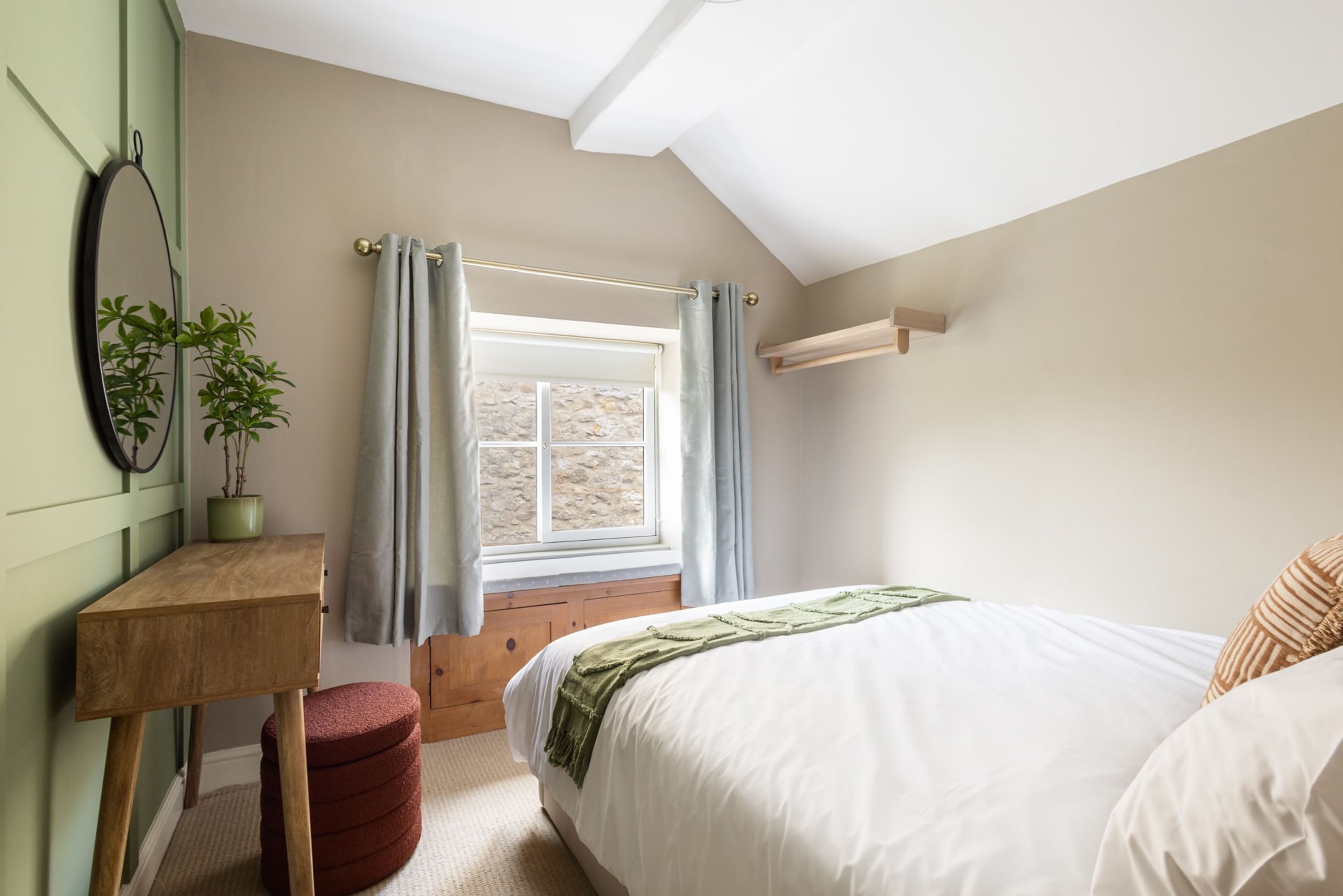 A bedroom with a bed, a window with curtains, a wooden bedside table, a red ottoman, and a green plant.