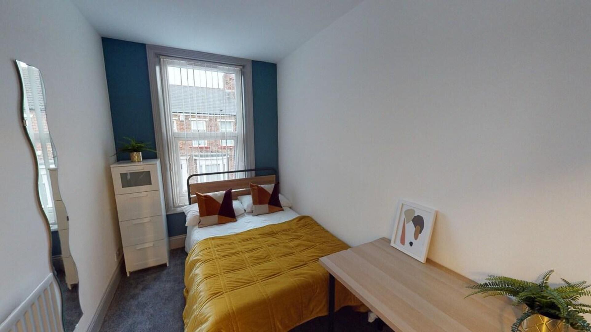 A bedroom with a bed, a bedside table, a desk, a window, and a mirror.