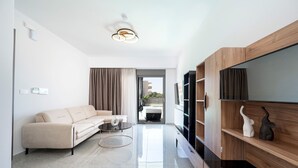 Un salon moderne avec un canapé beige, une table basse, un téléviseur à écran plat, des étagères en bois et une vue sur l’extérieur par une porte ouverte.
