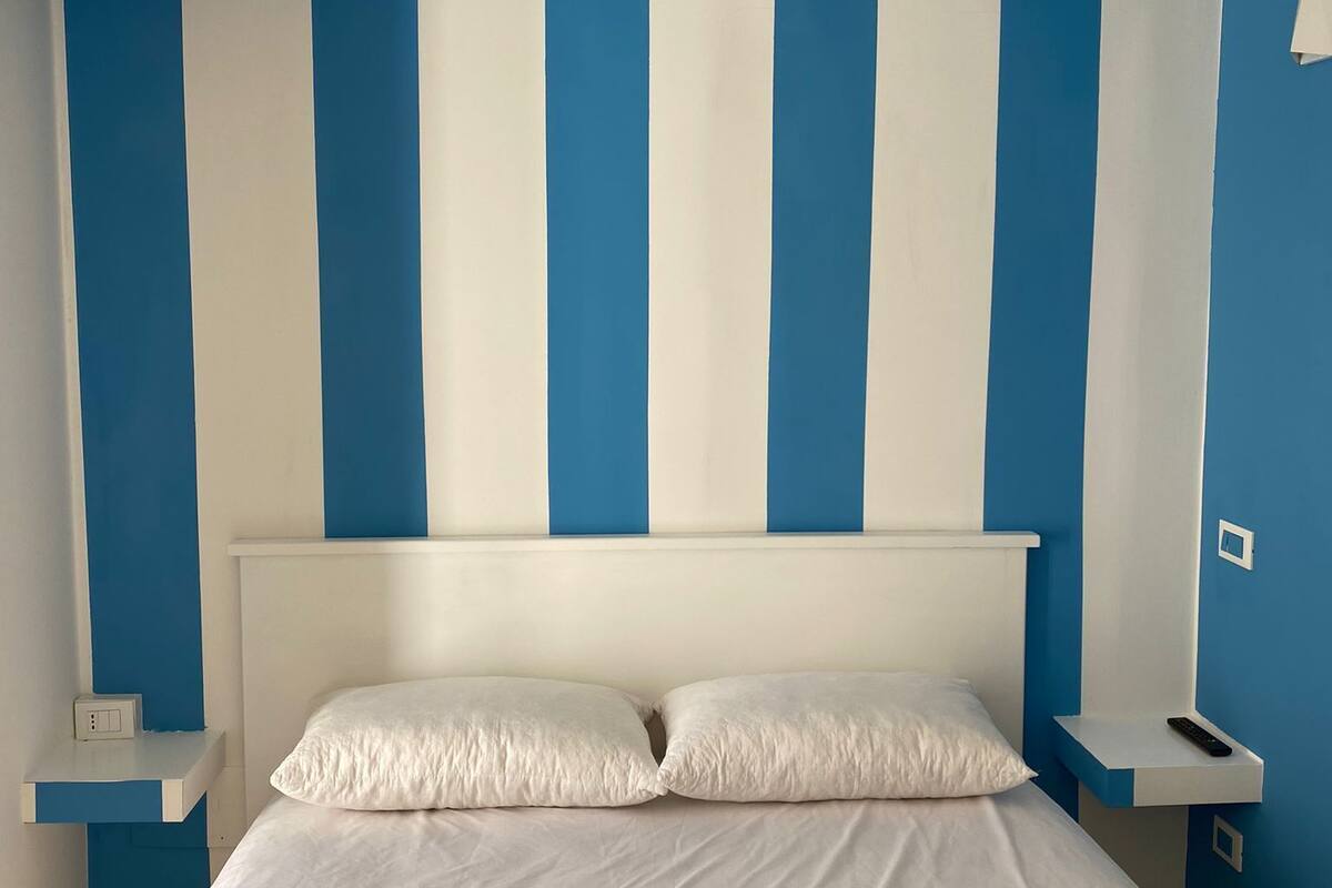 Un dormitorio con una cama blanca, paredes a rayas azules y blancas, y un cabecero blanco.