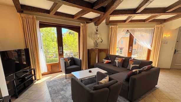 Een woonkamer met een bank, fauteuils, een salontafel, een televisie en grote ramen met gordijnen.